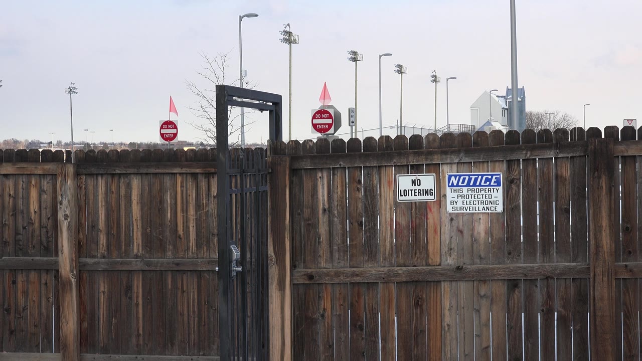In Westminster, Colorado, the frame stays on the brown fence and two posted warning signs, with the black gate near center and tall light poles rising behind it, no visible people.