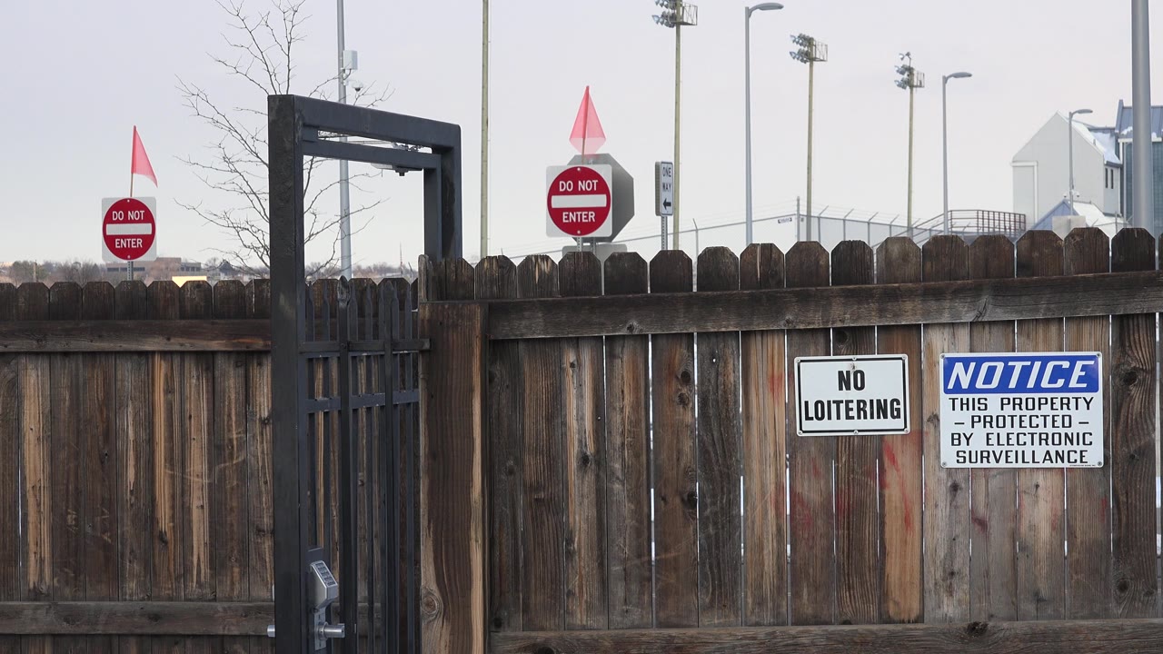 Weathered wooden fence in Westminster, Colorado with a black iron gate at left, no loitering and surveillance signs on the boards, snow at the base, and no visible people.