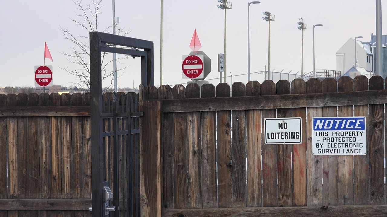 Westminster, Colorado fence view with the iron gate near the left side, security and no loitering signs centered on the wood, red traffic signs beyond the fence, and no visible people.