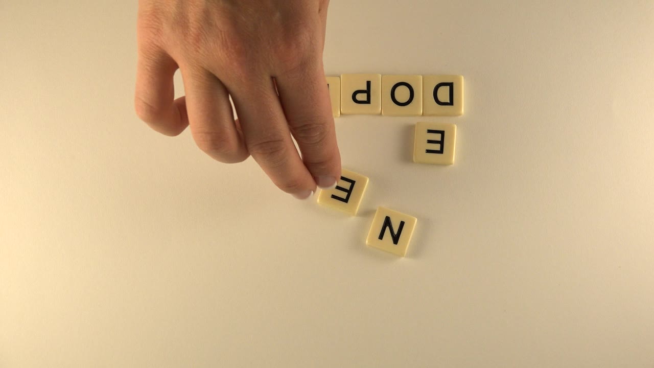 A hand hovers near a loose letter tile while other cream tiles sit partly arranged on the pale tabletop in Westminster, Colorado.
