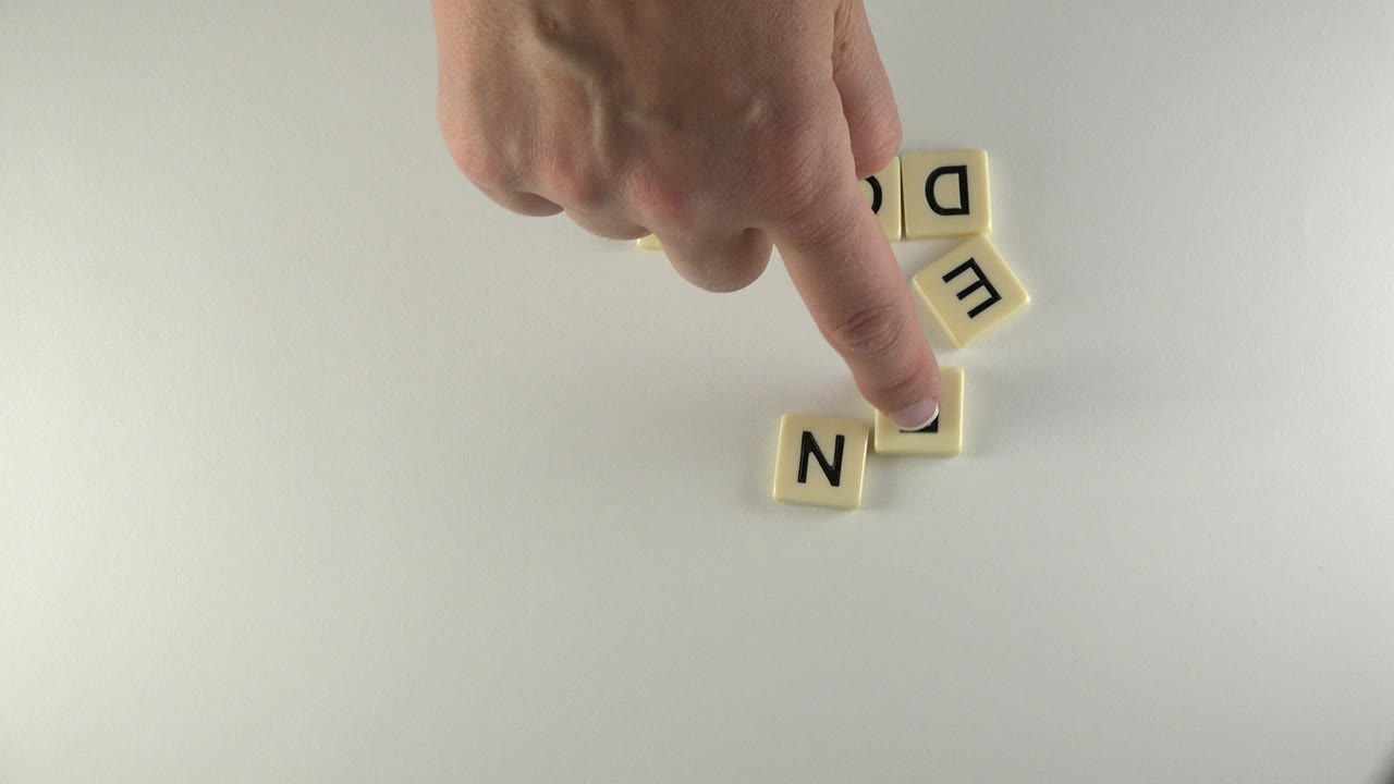 Hand presses a cream letter tile beside scattered letter tiles on a pale tabletop.