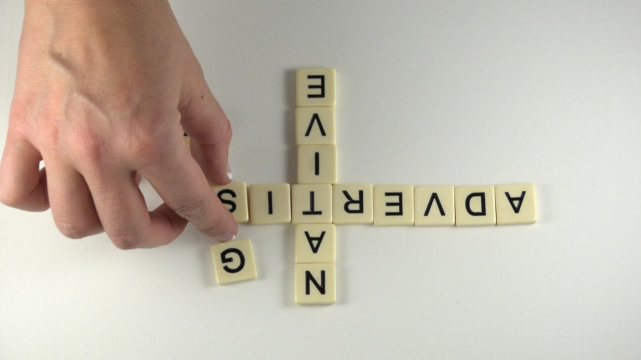 Hand near letter tiles spelling ADVERTISE and NATIVE on a white tabletop.
