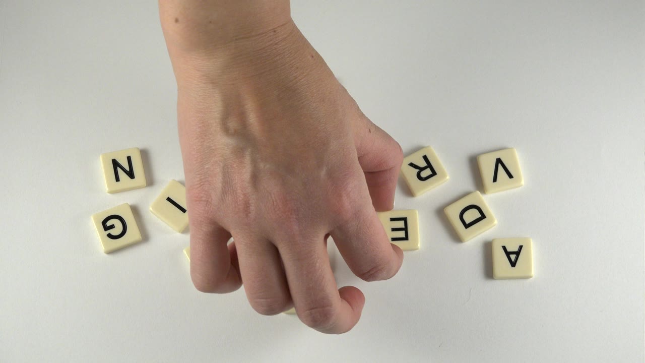 Hand above scattered cream letter tiles on a pale table.