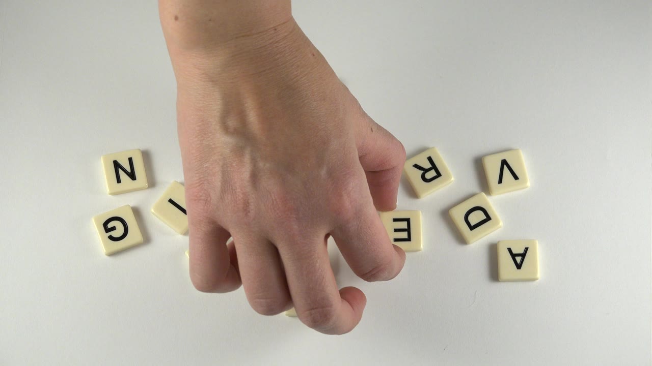 At 0:01 in Westminster, Colorado, the hand reaches lower into the frame while letter tiles remain scattered on both sides of the pale table.
