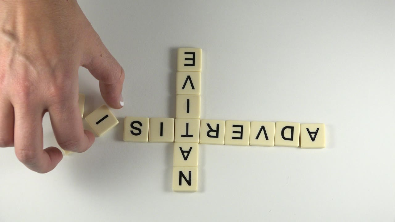 At 0:00 in Westminster, Colorado, a hand holds a cream letter tile near the left end of ADVERTISING while NATIVE crosses it vertically on a white tabletop.