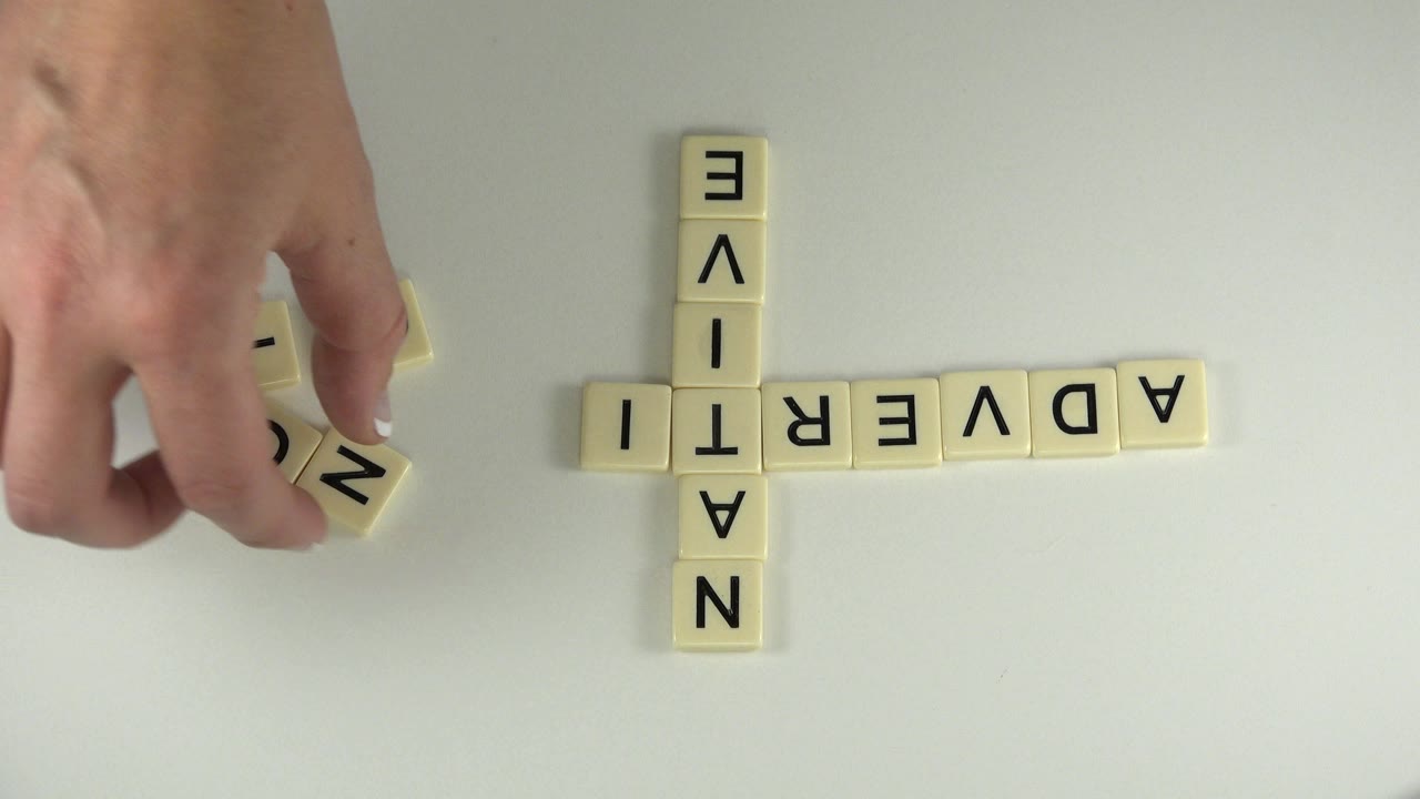 An adult hand reaches in over a white tabletop in Westminster, Colorado, with pale letter tiles already spelling NATIVE vertically and ADVERTI horizontally.