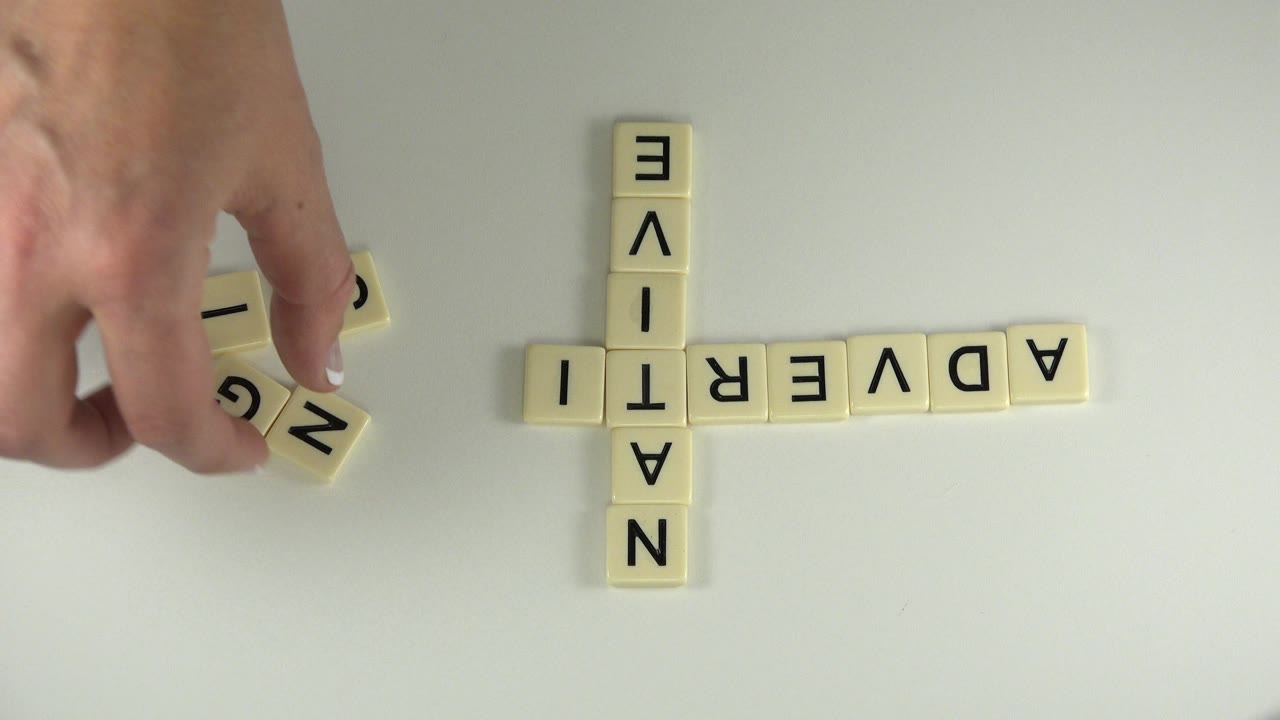 An adult hand holds loose letter tiles near the left side of the tabletop layout, with NATIVE vertical and ADVERTI horizontal visible in Westminster, Colorado.