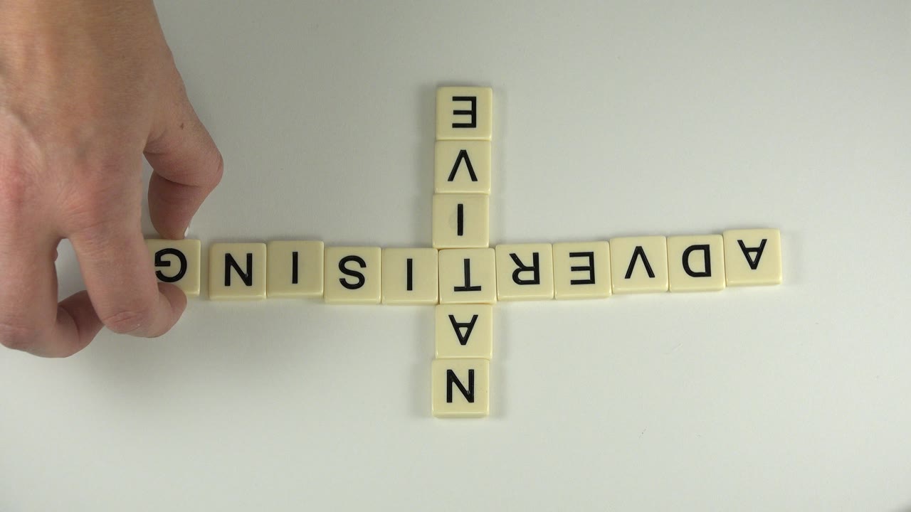 Hand touches cream letter tiles spelling ADVERTISING and NATIVE on a pale tabletop.