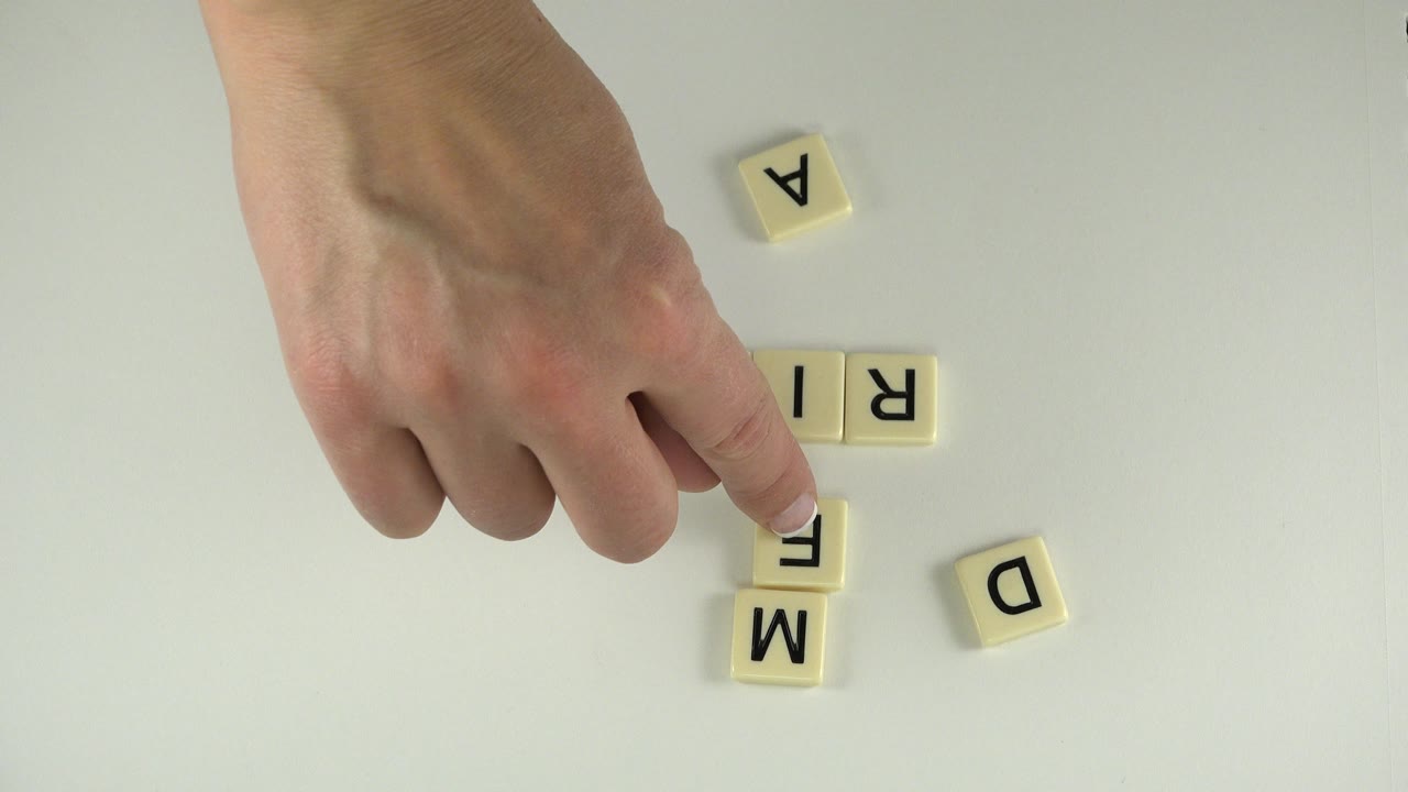 In Westminster, Colorado, the hand presses near a vertical tile arrangement while loose letter tiles sit around the pale tabletop.