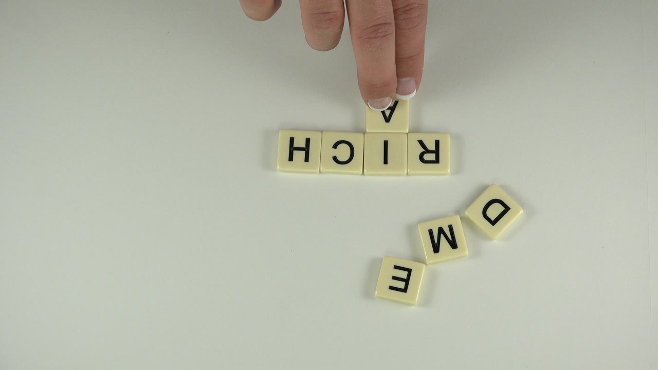A fingertip presses a cream letter tile above the RICH row while loose D, M, and E tiles sit below on the tabletop in Westminster, Colorado.