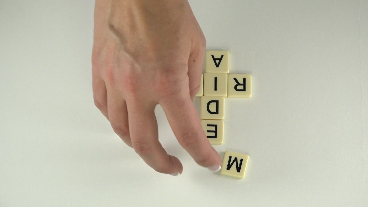 In Westminster, Colorado, a hand reaches toward a loose M tile near a crossword-style cluster of cream letter squares on a white surface, with no visible face.
