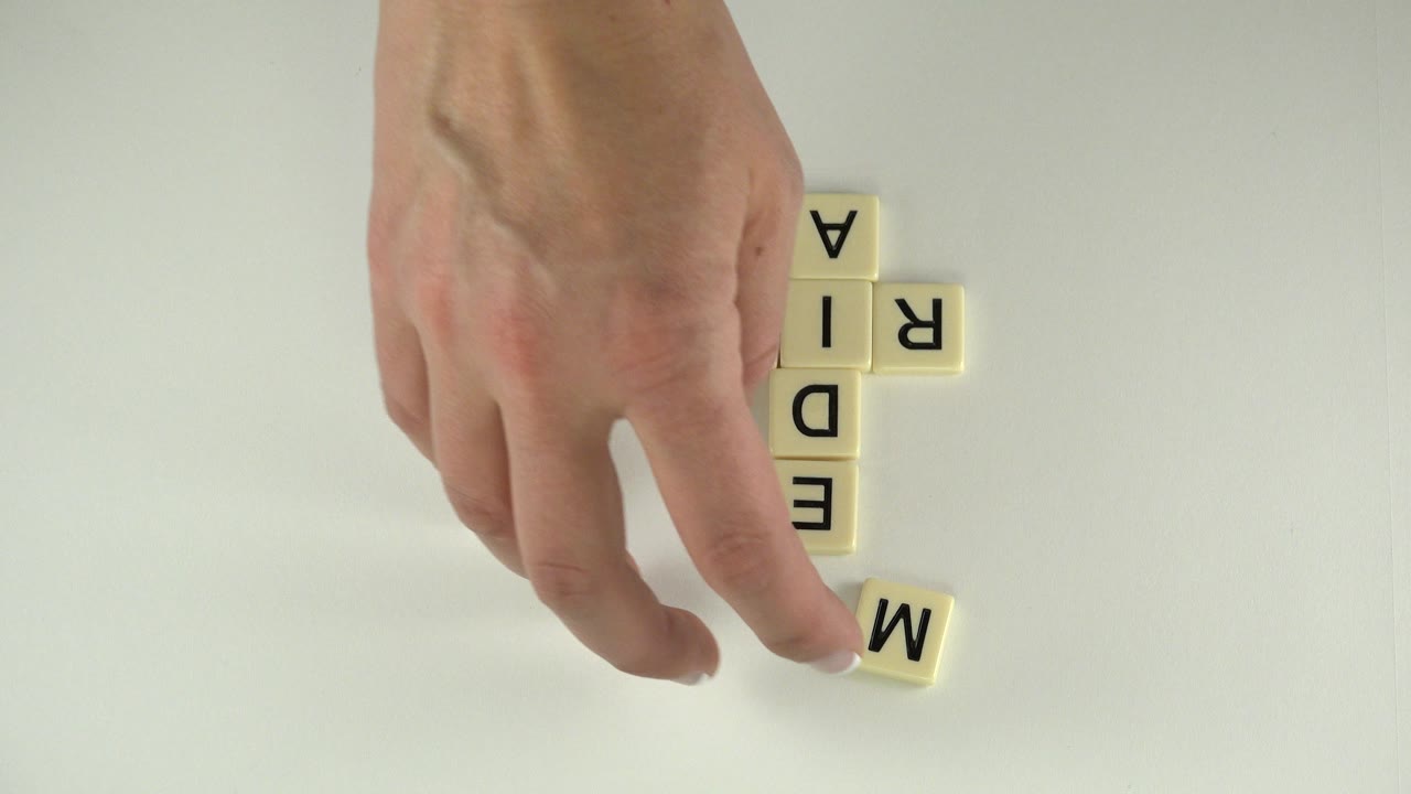 In Westminster, Colorado, the hand hovers over a loose M tile beside vertical MEDIA letters and a horizontal R tile on a white tabletop, with no visible face.