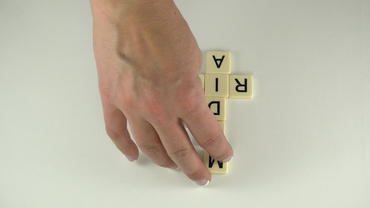 Hand placing the M tile beside cream crossword letters on white.
