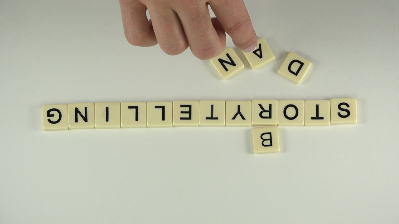 At 0:00 in Westminster, Colorado, a hand hovers over cream letter tiles while STORYTELLING is arranged horizontally and loose A, N, and D tiles sit above it.