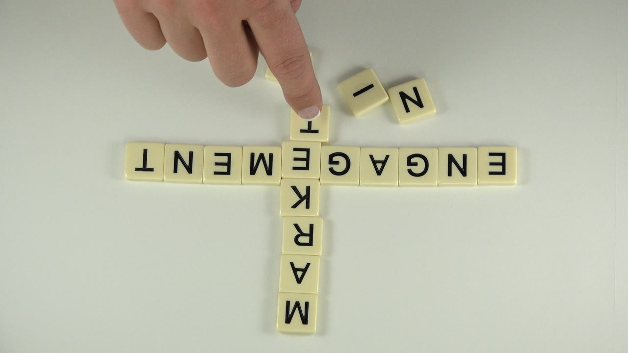 At 0:01 in Westminster, Colorado, the hand presses near a loose letter tile above the crossed engagement and marketing block words.