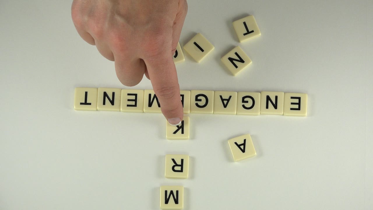 Hand pointing at letter tiles spelling ENGAGEMENT and MARK on a tabletop.