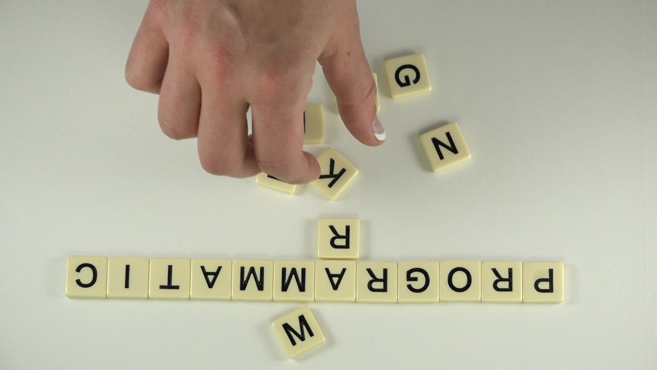 In Westminster, Colorado, the hand presses a loose letter tile near scattered G and N blocks while PROGRAMMATIC remains aligned below.