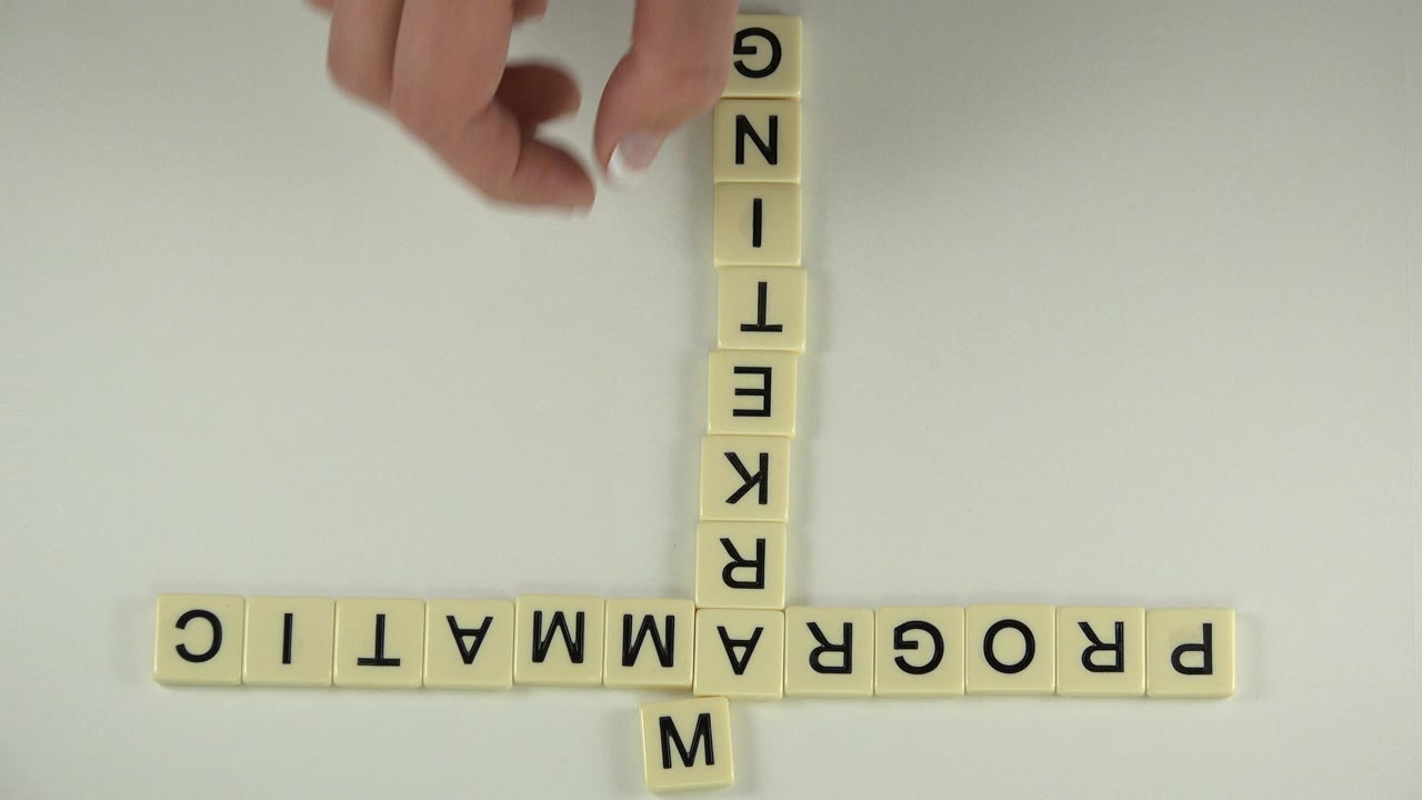 A manicured hand reaches in from the top edge toward a grid of cream letter tiles spelling PROGRAMMATIC across and MARKETING down on a pale tabletop.