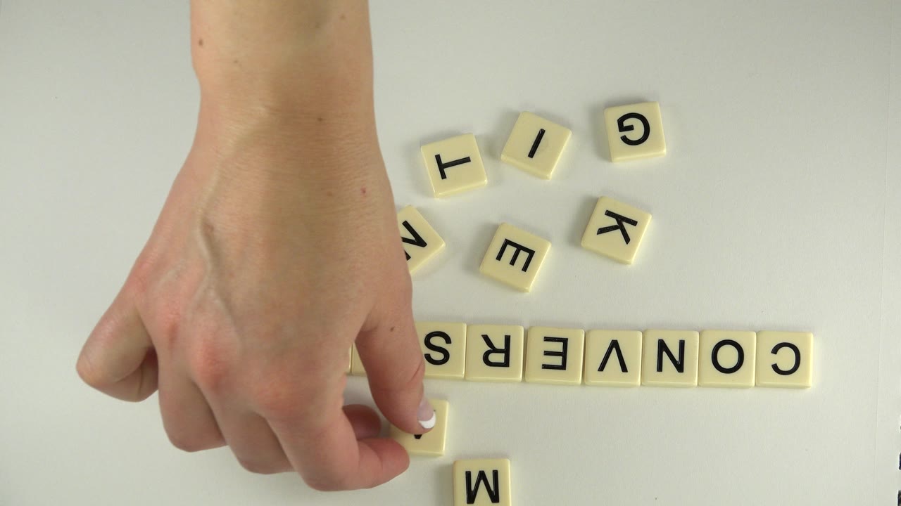 The hand remains near the left side of the overhead frame while loose cream tiles sit above the assembled word CONVERSATION on the pale surface.