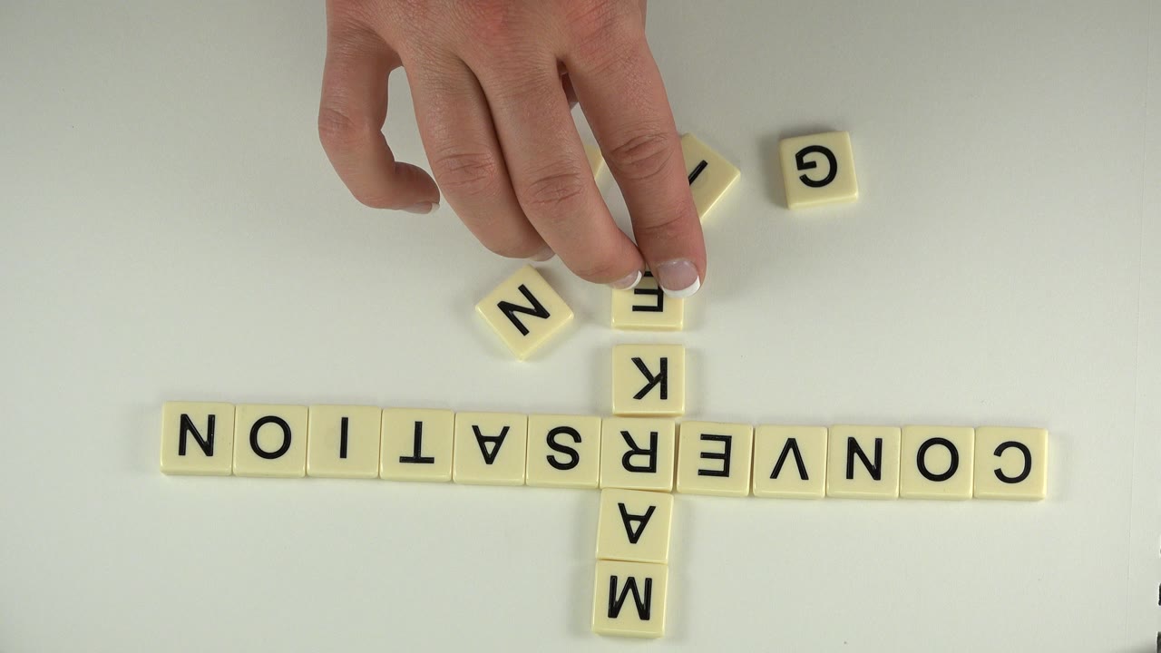 Hands place letter tiles beside a crossword-style word grid on a white table.