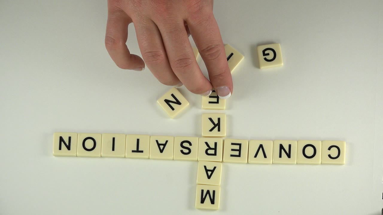 At one second, a hand positions a cream letter tile above the vertical MARKETING tiles, with loose N and G tiles nearby on the white table in Westminster, Colorado.