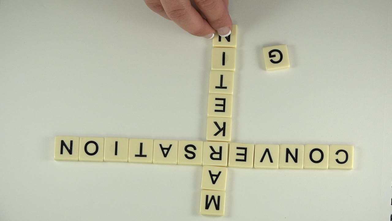 Inverted cream letter tiles on a pale tabletop spell CONVERSATION across and MARKETING vertically in Westminster, Colorado footage, with fingertips touching the top tile.