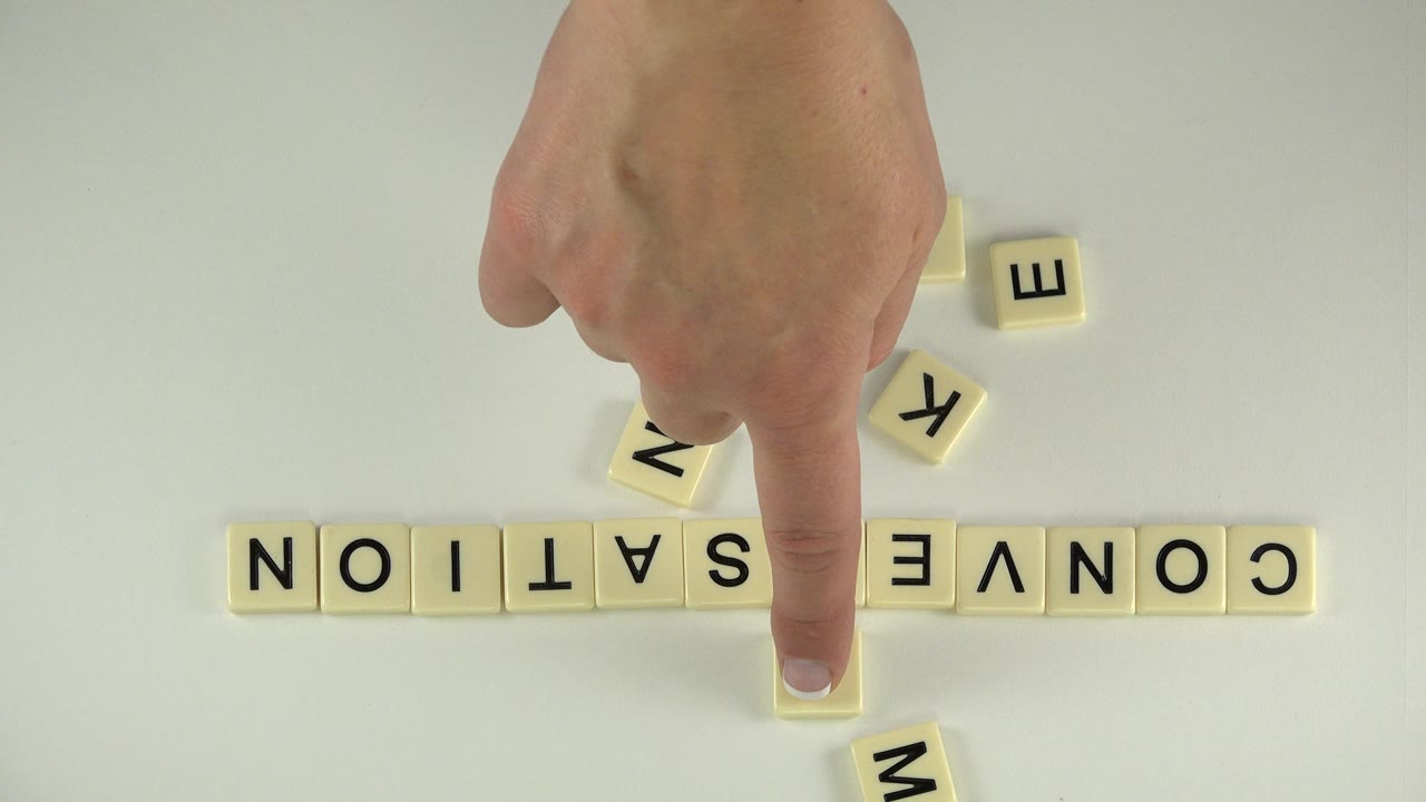 Hand points at a center tile among letter tiles spelling conversation.