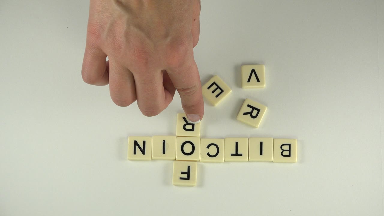 Hand points at letter tiles spelling BITCOIN and FOREVER on a tabletop.