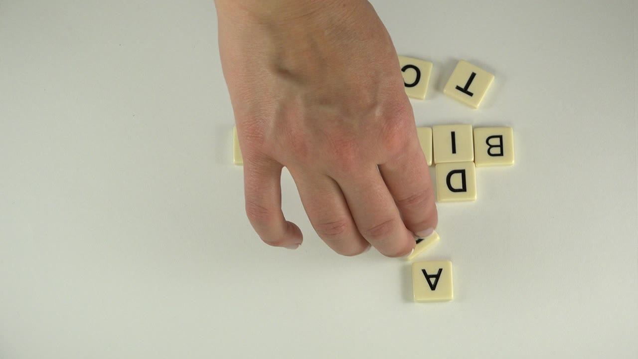 A hand hovers over the white table while several cream tiles form the early vertical structure of the word Addict in the Westminster, Colorado scene.