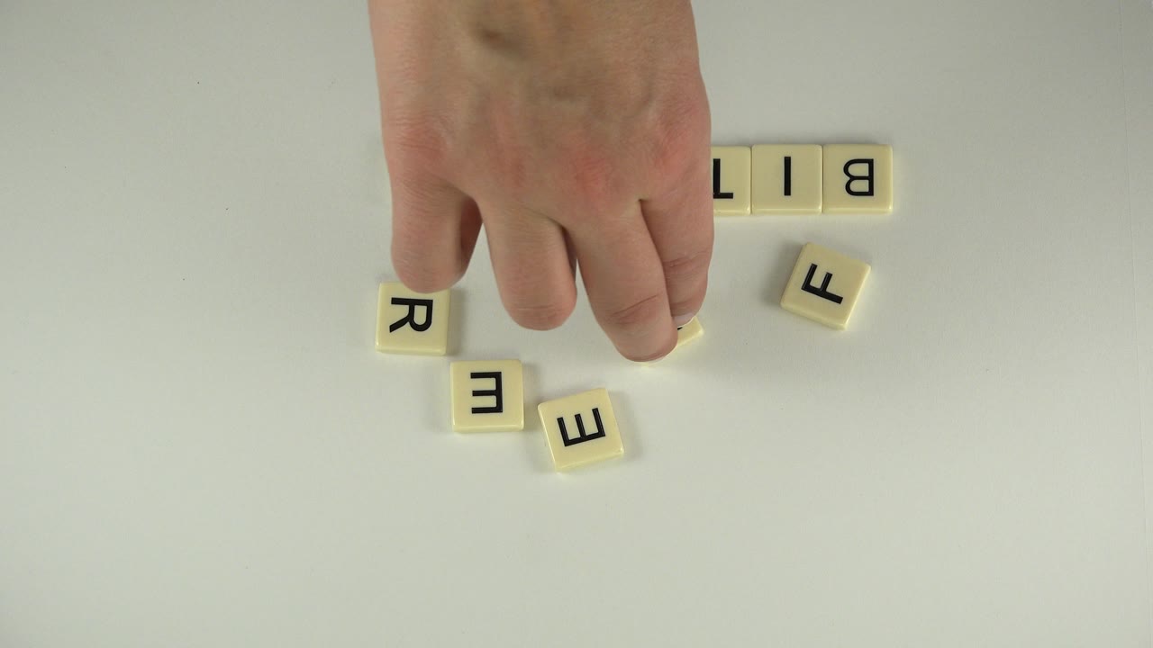At 0:00 in Westminster, Colorado, a hand reaches among cream letter tiles near the start of a tabletop spelling arrangement, with no visible face.