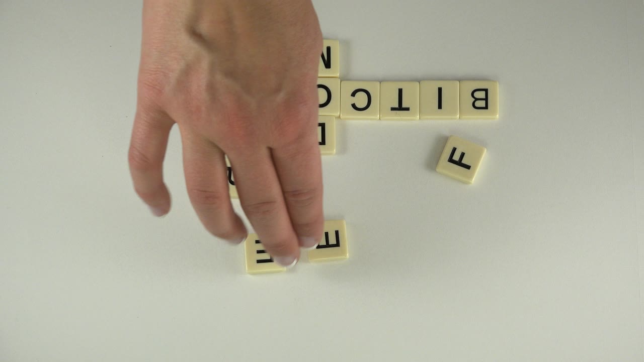 In Westminster, Colorado, the hand presses near two small letter tiles below the upside-down BITCOIN row, with a loose F tile set off to the right.