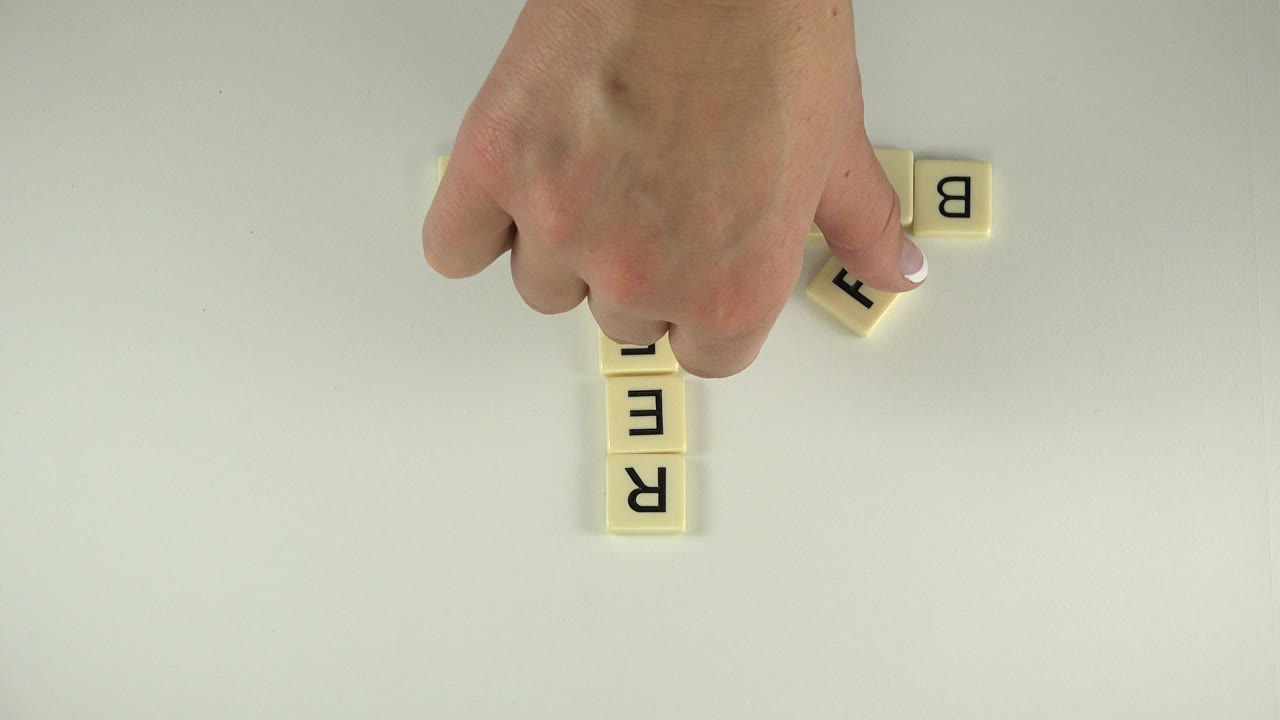 A hand hovers near upright and sideways letter tiles on the pale table as the word layout begins to expand.