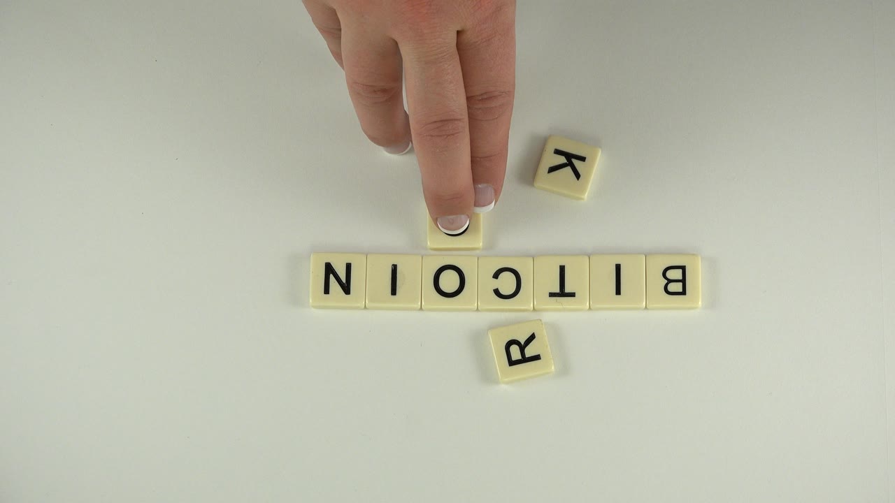 Hand placing a letter tile above the word BITCOIN on a pale tabletop.