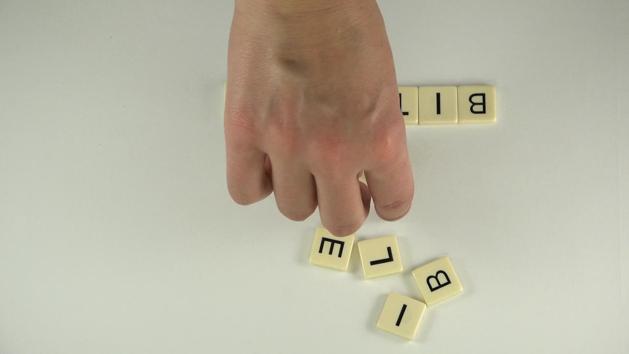Adult hand reaches over cream letter tiles on a pale tabletop in Westminster, Colorado, with partial BIT tiles visible near the top edge.