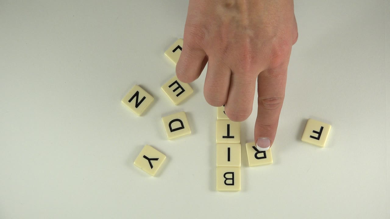A fingertip presses a cream tile beside a vertical stack of letters on a pale tabletop in Westminster, Colorado, with several loose tiles around it.