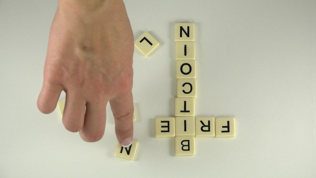 At 0:00 in Westminster, Colorado, an adult hand points toward a loose M tile while BITCOIN is spelled vertically and FRIEND is partly visible on a pale tabletop.