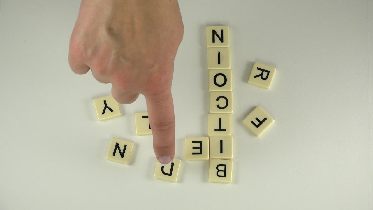 A finger presses near a single cream tile while the vertical BITCOIN word remains visible on the pale tabletop in Westminster, Colorado.