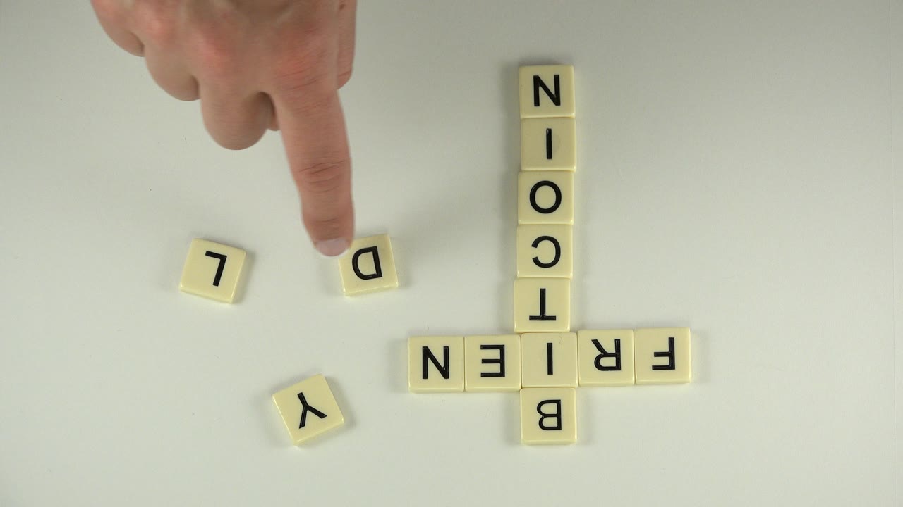 A finger hovers over a loose D cube beside the yellow tile words bitcoin and frien on a white tabletop in Westminster, Colorado.