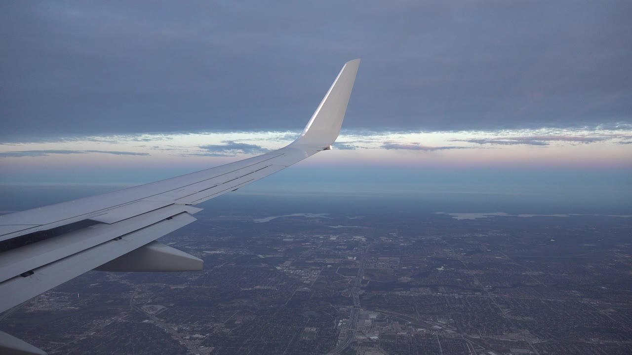 At the start of the Playa del Carmen, Mexico clip, a white airplane wing stretches across a cool blue-gray view of land, horizon, and layered cloud cover, with no visible people.