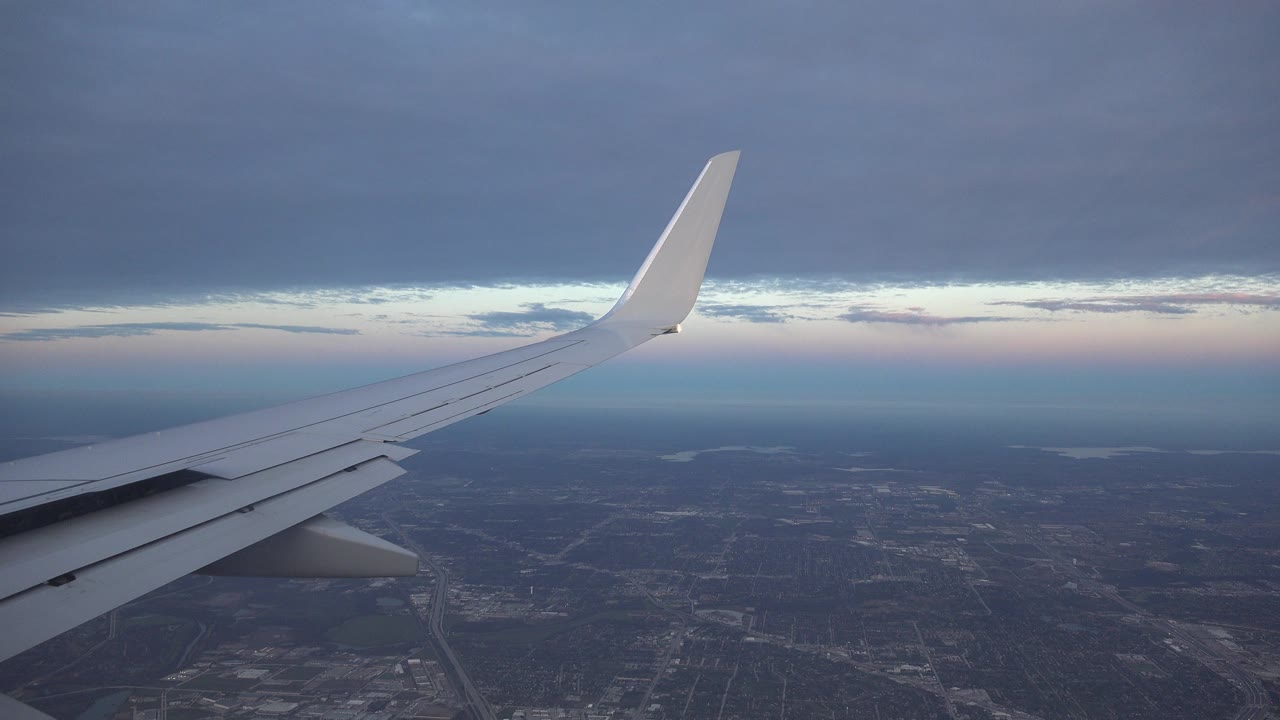 Opening frame from Playa Del Carmen, Mexico shows a white airplane wing stretching across a blue-gray sky over a dark city grid, with no visible people.