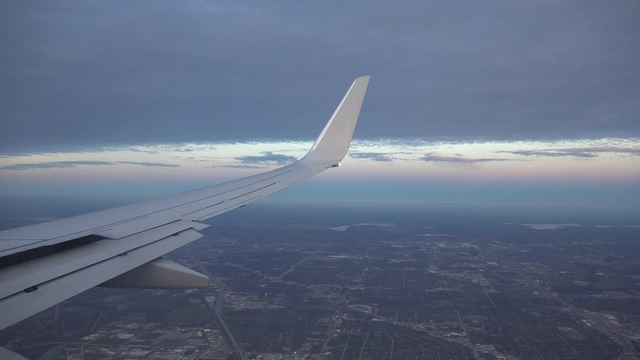 At 0:01, the Playa Del Carmen approach view holds on the wing as the city texture below becomes slightly clearer under a narrow pink sunset line, with no visible people.