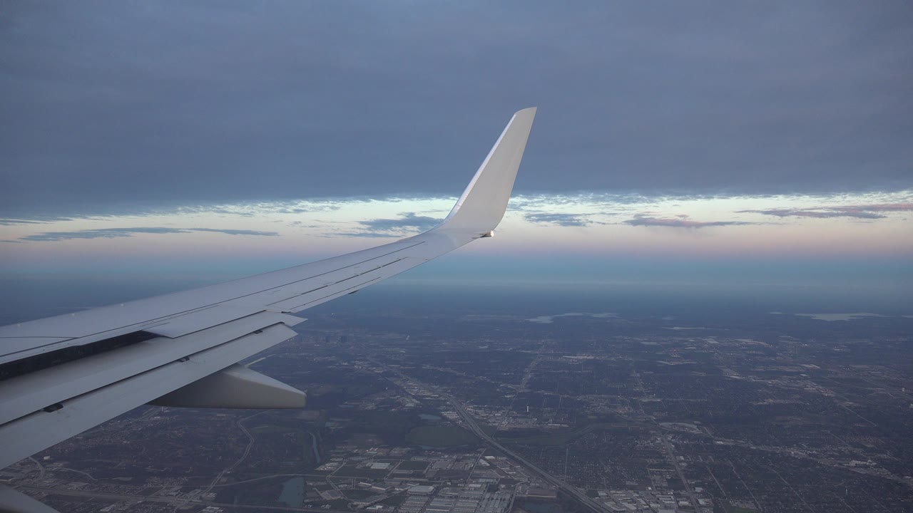 White airplane wing seen from a passenger window above Playa del Carmen, Mexico, with city terrain below, a pale horizon, dark cloud cover, and no visible people.