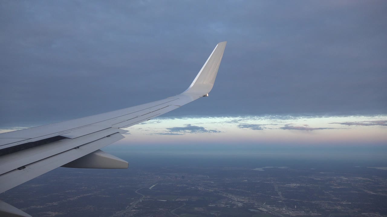 Passenger-window view over Playa del Carmen, Mexico with a white airplane wing crossing the frame, dark cloud cover above, distant land below, and no visible people.