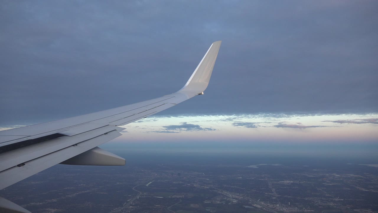 A white aircraft wing extends diagonally over the Playa del Carmen, Mexico area, with a blue-gray cloud ceiling, faint horizon light, distant land, and no visible people.