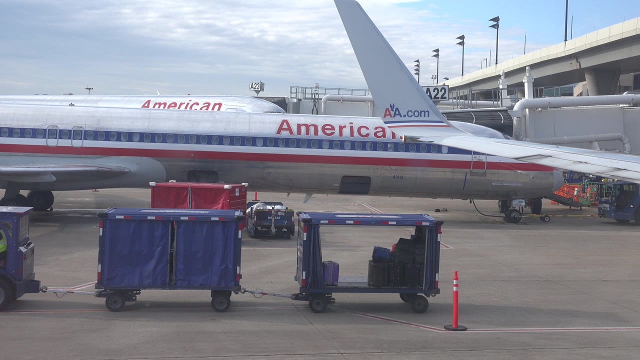 Silver American Airlines aircraft at a Playa Del Carmen Mexico airport gate with blue luggage carts, red cart, orange cone, jet bridge, bright clouded sky, and small airport workers near the service area.