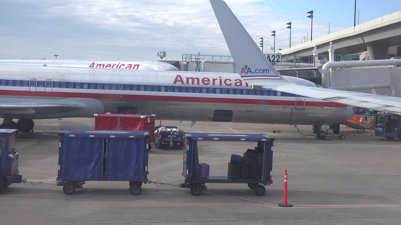 A blue luggage cart and a smaller loaded cart sit across the foreground while the silver American Airlines fuselage, wing, gate A22 sign, and jet bridge remain visible in Playa Del Carmen Mexico.