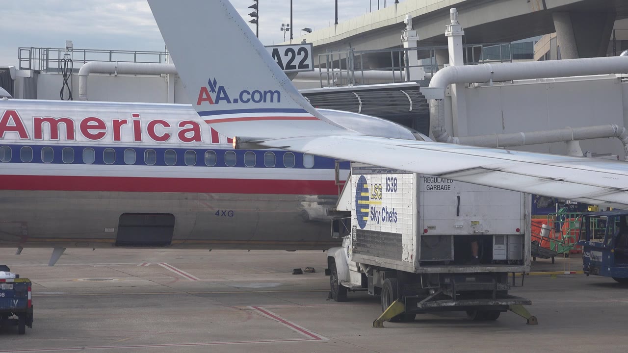 At 0:00, an American Airlines aircraft side and tail fill the frame in Playa Del Carmen, Mexico, with a Sky Chefs truck near the wing and no visible people.