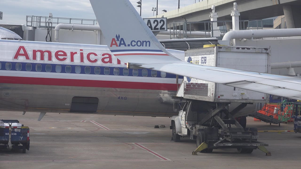 At one second, the Playa Del Carmen airport ramp view holds on the American Airlines fuselage, AA.com tail marking, white wing, attached jet bridge, and no visible people.