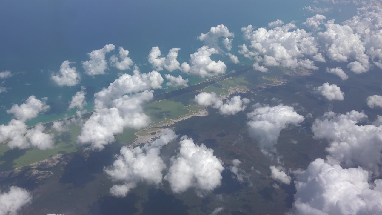 Aerial view near Playa Del Carmen, Mexico with scattered white clouds over blue water, green land, and darker ground below, no visible people.