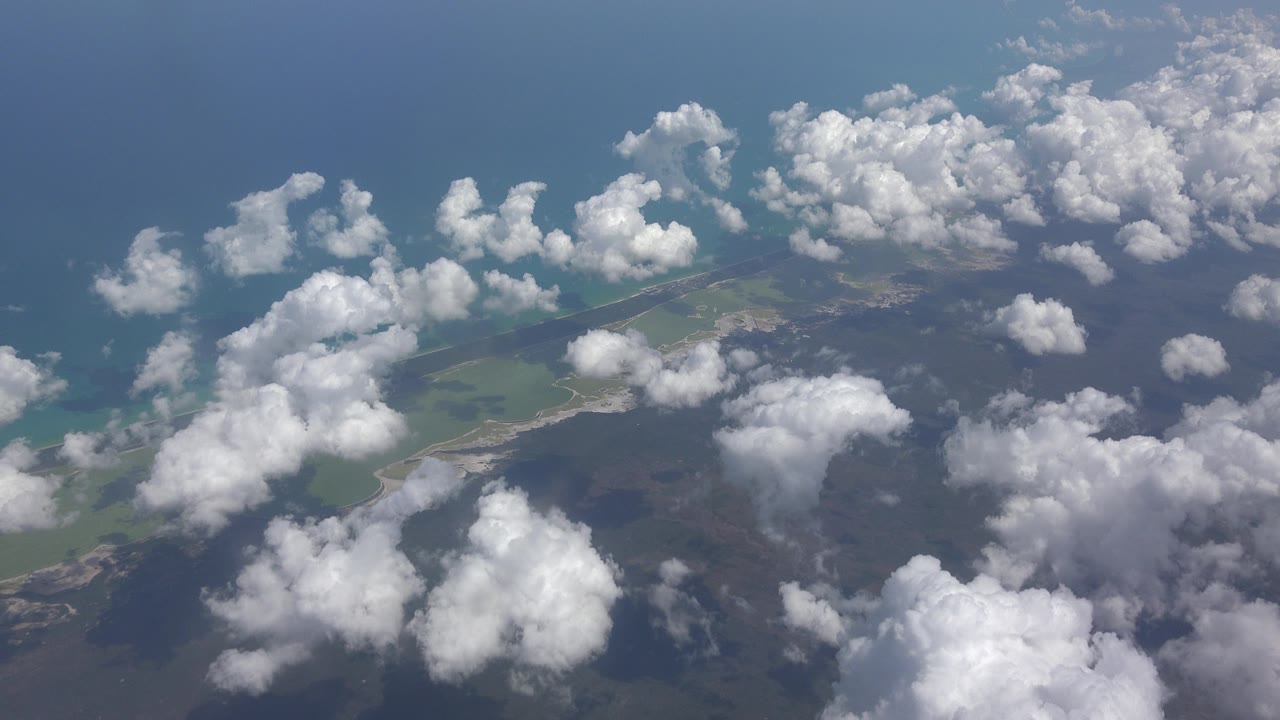 High view over Playa Del Carmen, Mexico, with scattered white clouds above blue water, pale beach edges, and green land below, with no visible people.